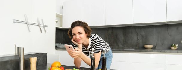 Nainen taivutus keittiössä laskuri älypuhelimella, etsivät terveellistä reseptiä verkossa matkapuhelimella, seisoo leikkuulauta ja vihannekset, ruoanlaitto ainesosat. - Valokuva, kuva