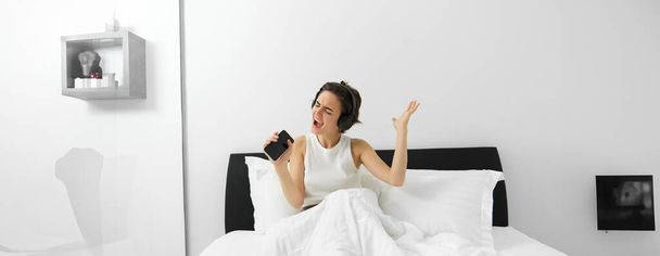 Image of modern, beautiful woman sitting on bed, singing into smartphone, listening music in wireless headphones. - Photo, image