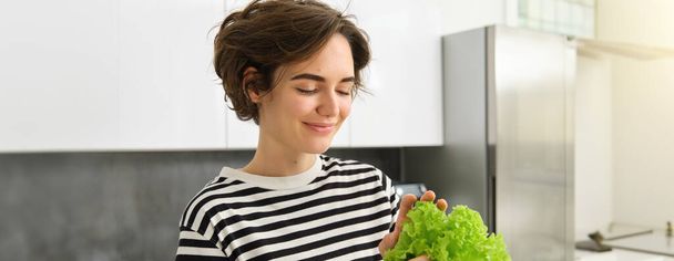 Portret van jonge glimlachende vrouw in de keuken, met verse groene sla bladeren, het bereiden van groenten voor salade, het koken van gezonde vegetarische maaltijd. - Foto, afbeelding