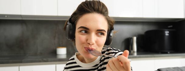 Close up portrait of young smiling woman in headphones, eating cereals with spoon and listening music, wearing earphones, sitting in the kitchen. - Foto, imagen