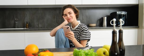 Portret van een jonge vrouw die online recepten zoekt met smartphone, naast groenten, saladeingrediënten en snijplank, glimlachend naar de camera. - Foto, afbeelding