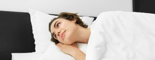 Close up portrait of smiling young woman, lying on white pillow, covered in linen sheets, looking aside at night table, waking up in hotel suit. - Фото, изображение
