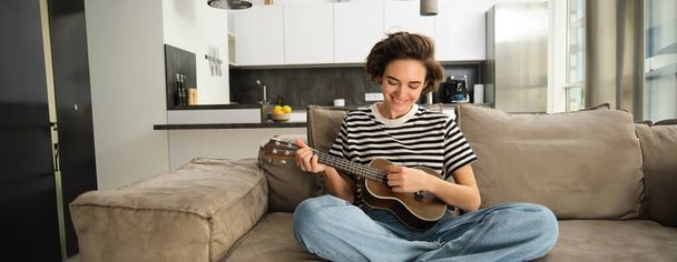 Retrato de una joven alegre tocando su ukelele, cantando y riendo, sentada en la sala de estar en casa. Concepto de estilo de vida y música - Foto, imagen