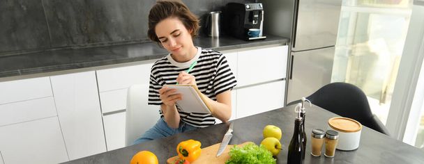 Junge brünette Frau schreibt Rezept auf, macht Notizen in Notizbuch, während sie Salat kocht, denkt an Einkaufsliste, sitzt in der Küche mit Gemüse auf dem Tisch. - Foto, Bild