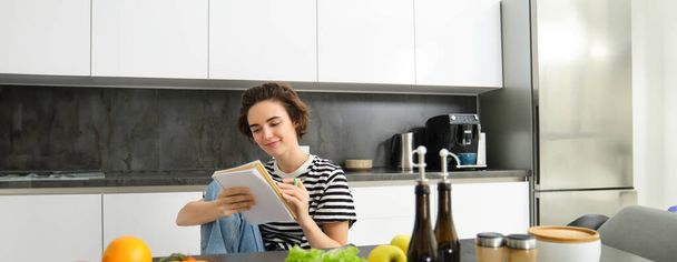 Retrato de una mujer joven cocinando, escribiendo notas, lista de comestibles en el cuaderno, creando una lista de comidas para cocinar a través de la comida, sentado en la cocina cerca de verduras y tabla de cortar. - Foto, Imagen