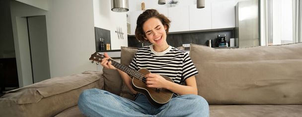 Πορτρέτο της χαρούμενης νεαρής γυναίκας που παίζει γιουκαλίλι, τραγουδά και γελάει, κάθεται στο σαλόνι στο σπίτι. Τρόπος ζωής και μουσική έννοια - Φωτογραφία, εικόνα
