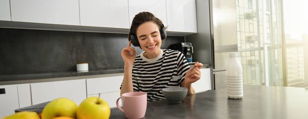 Portret van een gelukkige vrouw luistert graag naar muziek terwijl ze ontbijtgranen eet, draadloze koptelefoon draagt, glimlacht, in de keuken zit. - Foto, afbeelding