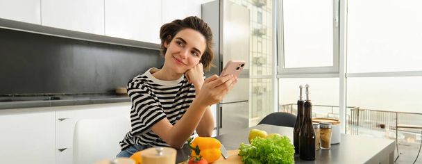 Retrato de chica morena cocinando comida en la cocina, buscando recetas en la aplicación de redes sociales, sosteniendo el teléfono móvil, de pie cerca de la tabla de cortar y verduras, preparando comida vegetariana saludable. - Foto, Imagen