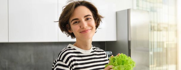 Primer plano retrato de mujer morena sonriente, de pie en la cocina, sosteniendo hoja de lechuga verde, mirando feliz a la cámara, comer alimentos saludables a dieta, hacer ensalada. - Foto, Imagen