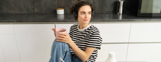 Retrato de mulher moderna elegante em fones de ouvido, ouve música e bebe chá, come cereais com leite para o almoço, senta-se na cozinha, olha para fora janela com sorriso relaxado. - Foto, Imagem
