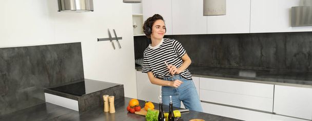 Glückliche Frau tanzt und kocht in der Küche, hört Musik über Kopfhörer, kocht eine Mahlzeit, bereitet Salat zu, schneidet Gemüse auf der Theke.. - Foto, Bild