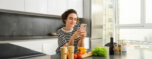 Porträt einer netten modernen Frau, die in der Küche kocht, Smartphone benutzt, Rezepte liest, Mahlzeiten für das Frühstück sucht, sich an Gemüsetheke und Schneidebrett lehnt. - Foto, Bild