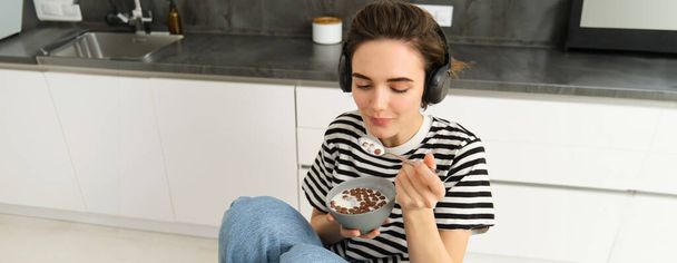 Szczęśliwa brunetka w kuchni, jedząca płatki z mlekiem na śniadanie, słuchająca muzyki w słuchawkach, jedząca poranny posiłek. - Zdjęcie, obraz