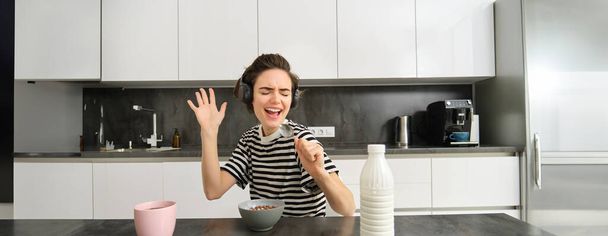 Glückliche und emotionale junge Frau, singend beim Frühstück, Müsli mit Milch, Musik über drahtlose Kopfhörer hörend, in der Küche sitzend. - Foto, Bild
