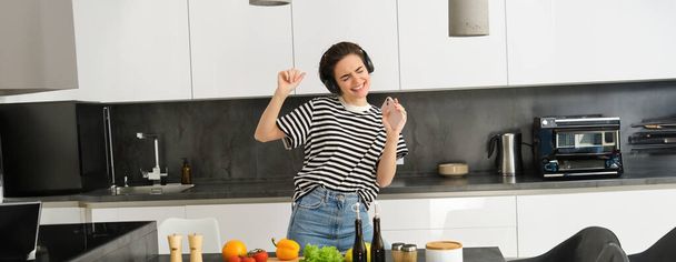Ξέγνοιαστη μοντέρνα γυναίκα με ασύρματα ακουστικά, ακούει μουσική, χορεύει και μαγειρεύει σαλάτα, ετοιμάζει φαγητό στην κουζίνα, τραγουδάει σε smartphone. - Φωτογραφία, εικόνα