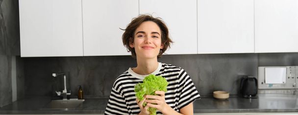Porträt einer glücklichen und gesunden jungen Frau, die ihrer Ernährung folgt, mit Salatblatt posiert und lächelt, in der Küche kocht, Vegetarierin liebt ihr Gemüse. - Foto, Bild