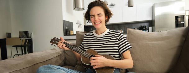 Unbekümmertes junges Model, braune Konzert-Ukulele spielend, mit Leidenschaft singend, mit kleiner Gitarre in der Hand auf Sofa sitzend. - Foto, Bild