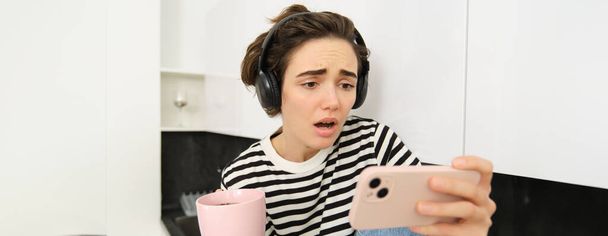 Portrait of woman looking with sympathy and sad face at smartphone screen, watching video on mobile phone app, drinking tea or coffee in the kitchen, frowning and looking with pity. - Photo, Image