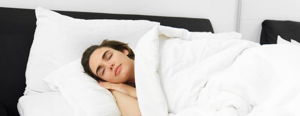 Bild der schönen jungen Frau, die auf einem bequemen Bett schläft, bedeckt mit weichem weißen Leintuch, die Augen geschlossen, das Gesicht ist ruhig und unbehelligt. - Foto, Bild