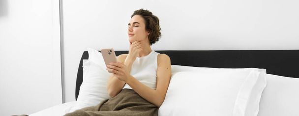 Retrato de mujer joven con los ojos cerrados y sonrisa complacida, sosteniendo teléfono inteligente, acostado y descansando en la cama, usando teléfono móvil. - Foto, Imagen