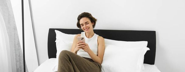 Portré aranyos, mosolygós fiatal nő a hálószobában, feküdt az ágyban okostelefonnal, nézte a mobil képernyőn boldog arc. - Fotó, kép