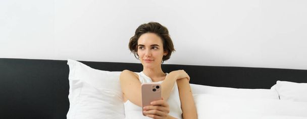 Πορτρέτο της χαμογελαστής μελαχρινής γυναίκας που αναπαύεται στην κρεβατοκάμαρά της, χρησιμοποιεί το κινητό της τηλέφωνο και ξαπλώνει στο κρεβάτι φορώντας τα ρούχα του σπιτιού της, ανταλλάσσοντας μηνύματα, στέλνοντας μηνύματα στην εφαρμογή smartphone. - Φωτογραφία, εικόνα