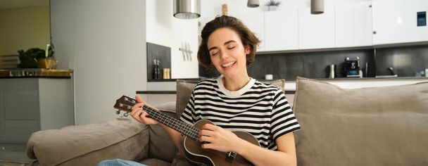 Bella giovane donna suonare ukulele, cantare e godersi il suo strumento musicale, trascorrere del tempo da sola a casa, rilassarsi in soggiorno. - Foto, immagini