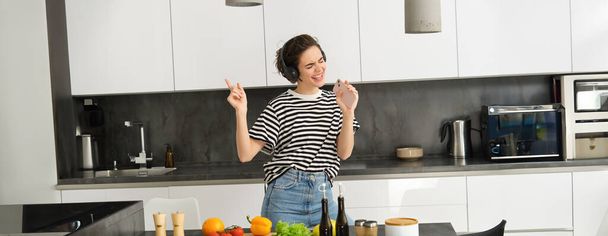 Portrait de fille heureuse cuisine salade et écouter de la musique dans les écouteurs, danser tout en faisant un repas dans la cuisine. - Photo, image