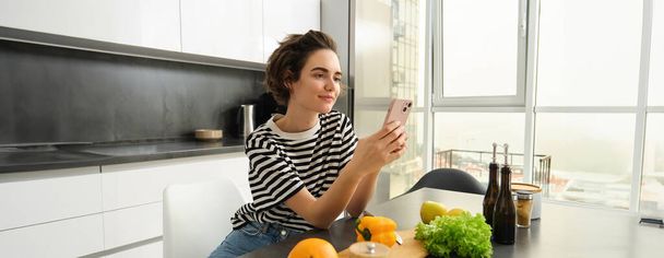 Portrait d'une fille brune cuisinant des aliments dans la cuisine, cherchant des recettes sur l'application de médias sociaux, tenant son téléphone portable, se tenant près de la planche à découper et des légumes, préparant un repas végétarien sain. - Photo, image