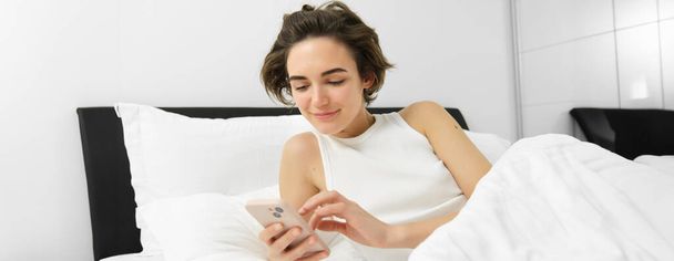 Afbeelding van vrouwelijk model in bed, kijken naar smartphone, verzenden van een bericht, zet wekker op mobiele telefoon, gaat slapen in haar slaapkamer. - Foto, afbeelding