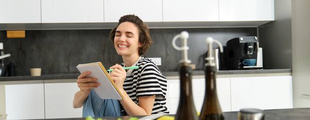 Zbliżenie szczęśliwej, uśmiechniętej młodej kobiety piszącej w notatniku, tworzącej przepis, listę posiłków na ten tydzień, siedzącej w pobliżu warzyw i oliwy z oliwek, gotującej w kuchni. - Zdjęcie, obraz