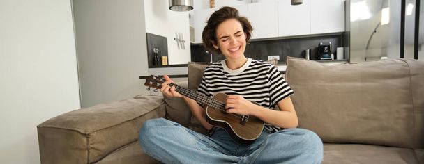 Młoda kobieta gra na ukulele z pasją, śpiewa piosenki, struganie struny, siedzi na kanapie w salonie w domu. Koncepcja stylu życia i muzyki - Zdjęcie, obraz