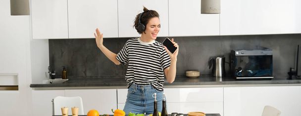 Portrait de femme dansante heureuse, cuisine dans la cuisine et écouter de la musique dans les écouteurs, hacher des légumes pour la salade et chanter avec un visage insouciant. - Photo, image