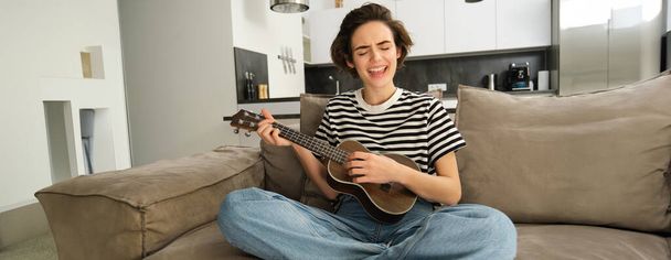 若い幸せな女性はソファーに座ってウクレレを演奏し,歌い,新しい楽器を学ぶことを楽しんでいます. - 写真・画像
