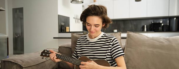 Portret wesołej młodej kobiety grającej na ukulele, śpiewającej i śmiejącej się, siedzącej w salonie w domu. Koncepcja stylu życia i muzyki - Zdjęcie, obraz