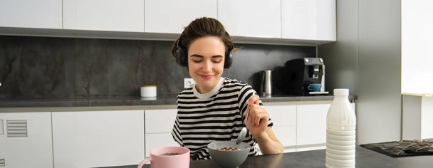 Femme moderne mignonne, étudiant petit déjeuner rapide, manger des céréales avec du lait et du café, écouter du podcast ou de la musique dans un casque sans fil. - Photo, image