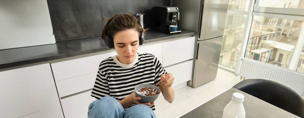 Jonge vrouw die 's morgens ontbijtgranen eet, studente die ontbijt, muziek luistert of podcast in draadloze koptelefoon. - Foto, afbeelding
