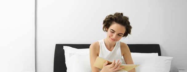 Πορτρέτο της γυναίκας που κάθεται στο κρεβάτι με ημερολόγιο, γράφει στο ημερολόγιο, σκέψεις ή συναισθήματα το πρωί, κρατώντας σημειωματάριο και χαμογελώντας. - Φωτογραφία, εικόνα