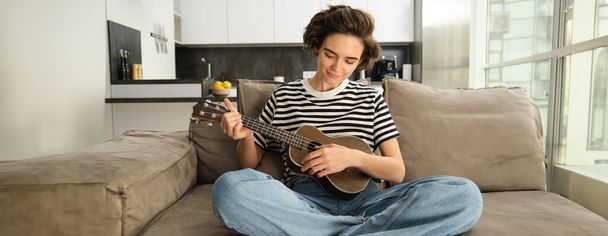 Yaşam tarzı ve hobi konsepti. Kanepede gülümseyen genç bir kadın, ukulele çalıyor, en sevdiği şarkı için tıngırdatıyor ve öğreniyor.. - Fotoğraf, Görsel