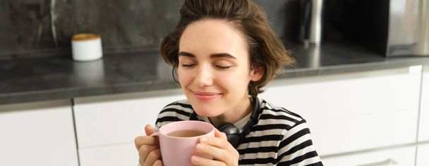 Gros plan portrait de femme aux yeux fermés et sourire heureux, boit du thé chaud avec satisfaction, assis dans une cuisine confortable. - Photo, image