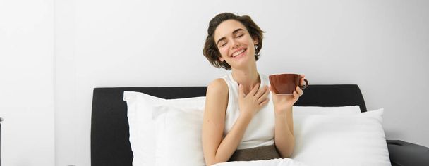 コーヒーを飲むケアフリーの女性の肖像画,朝のカップでベッドに座って,一日の始まりを楽しんで,目覚め,微笑んでいる. - 写真・画像