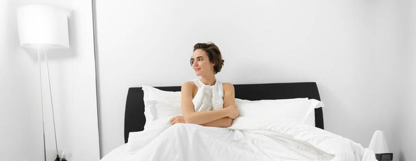 Πορτρέτο της νεαρής χαμογελαστής γυναίκας με τα ακατάστατα μαλλιά, που κάθεται στο κρεβάτι της, καλυμμένη με λευκά λινά σεντόνια, κοιτάζοντας στην άκρη με ονειρεμένη έκφραση προσώπου, ξυπνώντας το πρωί. - Φωτογραφία, εικόνα