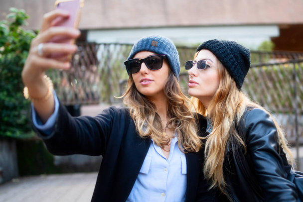Two fashionable women wearing beanies and sunglasses taking a selfie on a sunny day, capturing a moment of friendship. - Photo, Image