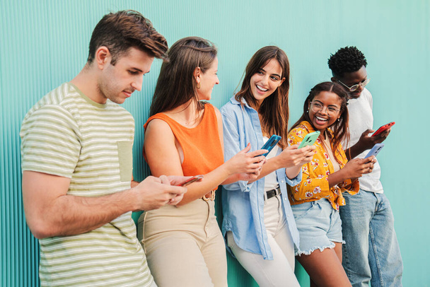 Multiracial group of happy teenage friends enjoying and smiling using their cell phone app at teal blue wall background. Diverse young student people having fun watching content on the social media - Photo, Image