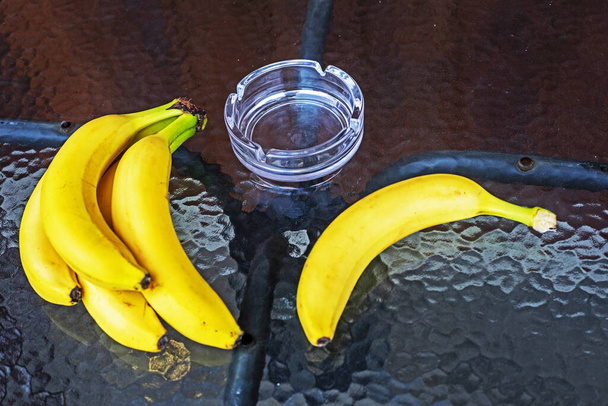 куча бананов и банан на стеклянном круглом столе в кафе вместе с пепельницей. зона для курящих - Фото, изображение