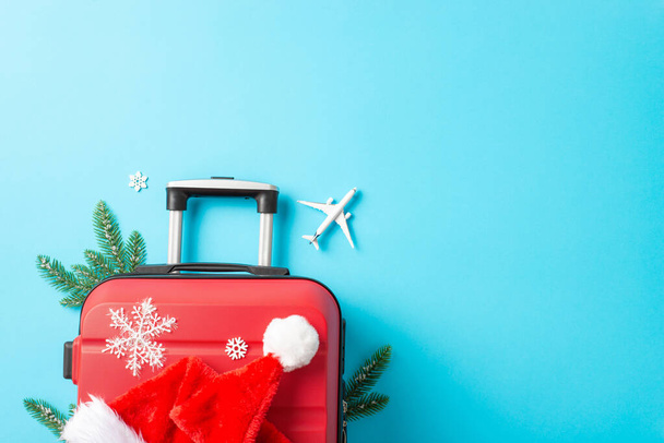 De kerstman is onderweg naar je feest. Boven foto van een rode koffer, klein vliegtuig, Santa 's hoed, sparren takken, sneeuwvlokken op een levendig blauw oppervlak voor tekst - Foto, afbeelding