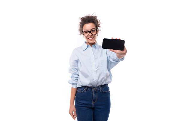 smukła młoda brunetka kręcone biznesu kobieta ubrana w jasnoniebieską koszulę pokazuje ekran smartfona. - Zdjęcie, obraz