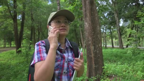 森林公園の電話で話しているカジュアルなドレスの生態学者の女性. 自然環境に影響を与える要因による研究結果の報告. 持続可能な自然保護への意識. - 映像、動画