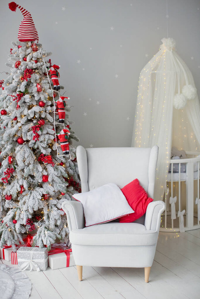 Χριστουγεννιάτικο δέντρο με κόκκινες διακοσμήσεις, λευκή πολυθρόνα με μαξιλάρια και κούνια μωρού με θόλο σε χαριτωμένο παιδικό δωμάτιο εσωτερικό με γκρι τοίχους - Φωτογραφία, εικόνα