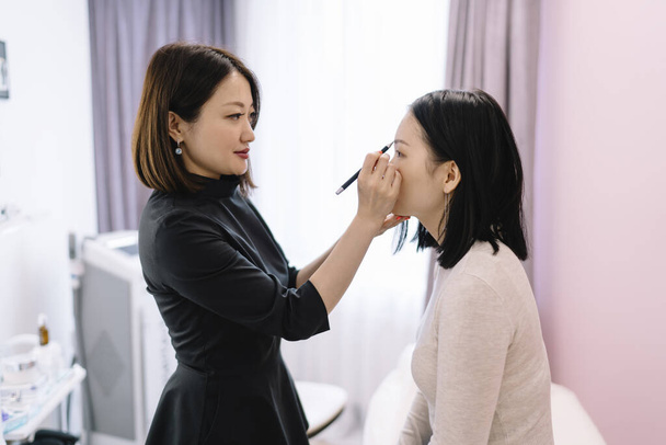 Side view of concentrated young Asian female beauty specialist in black uniform doing makeup with pencil to client while working in modern salon - Zdjęcie, obraz